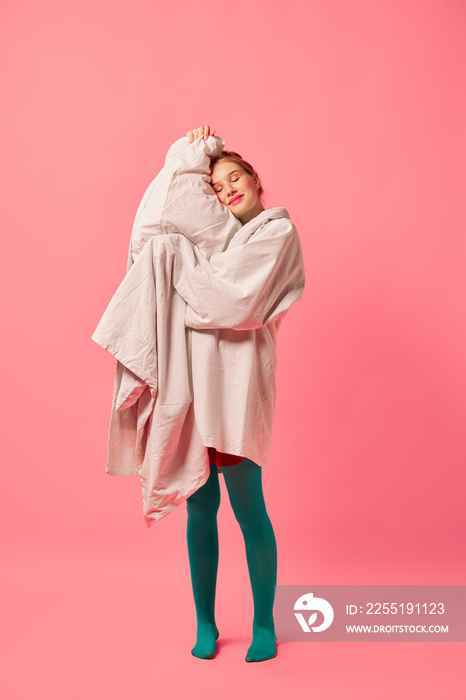 Beautiful young girl wrapped in soft blanket standing isolated over pink background. Comfrort, fashion, art, creativity