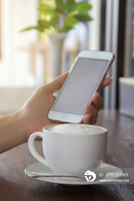 Woman’s hand holding a smart phone and a cup of hot coffee on the table.