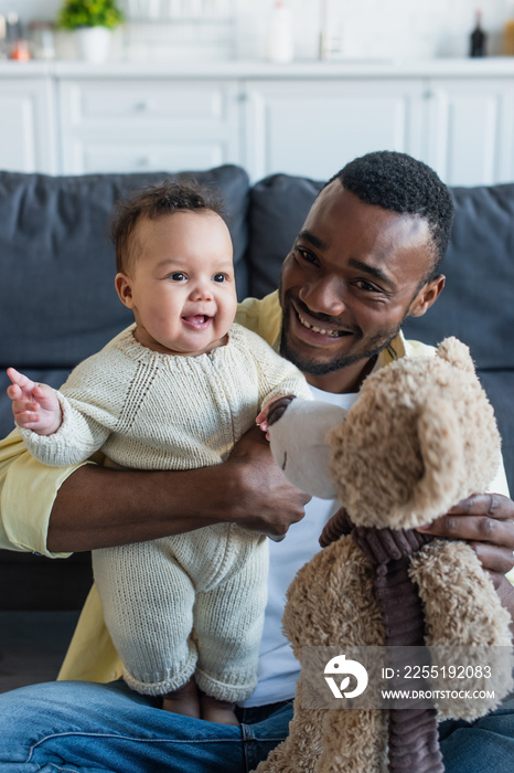 happy african american man holding teddy bear near cheerful baby