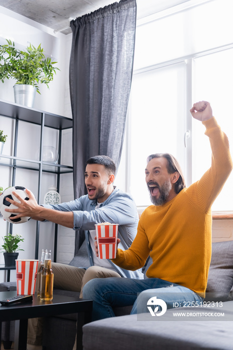 Excited man showing win gesture while watching football championship with Hispanic son at home