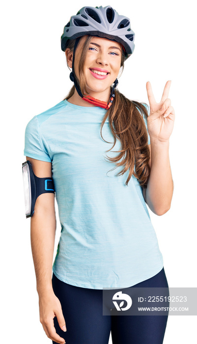 Beautiful caucasian young woman wearing bike helmet smiling with happy face winking at the camera doing victory sign. number two.