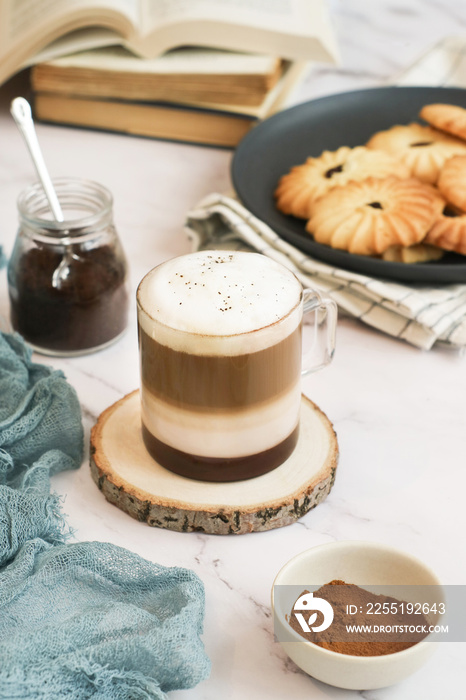 An angle view of a cup of layered macchiato coffee topped with foam with books and plate of cookies in background. A drink is based on the espresso and steamed milk combination. It’s similar to latte