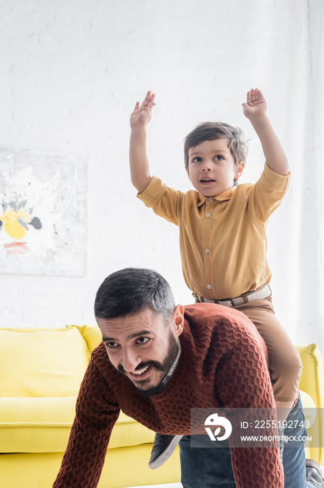 Cheerful boy sitting on back of hispanic grandfather at home, two generations of men