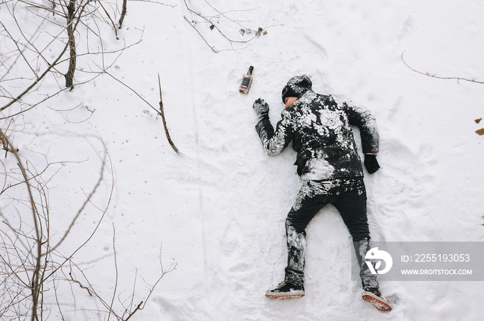 A drunken homeless and dirty man in a black coat lies, sleeps on his stomach on white snow in a frosty winter with a bottle of alcohol, strong whiskey. Photography, copy space.