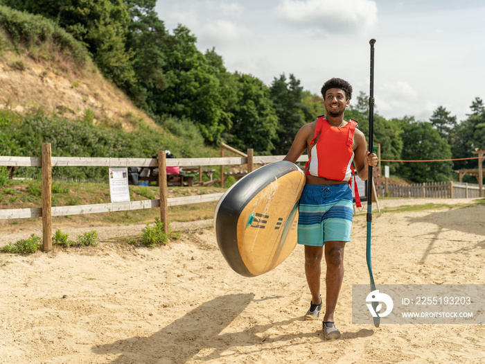 Young man carrying paddleboard and oar on beach