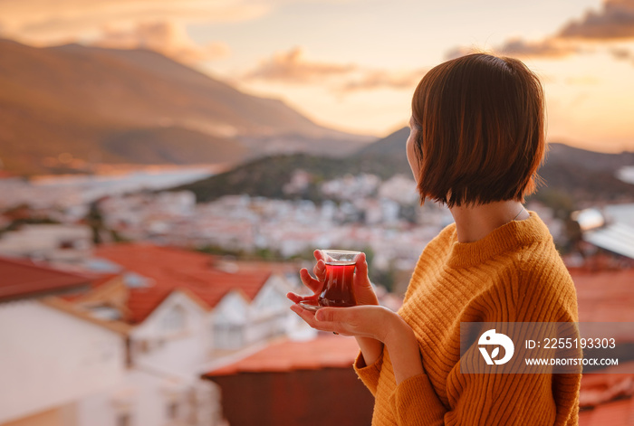 Woman drinking turkish tea from traditional turkish teacup and enjoys panorama over sunset of Kas resort town of Mediterranean sea in Turkey