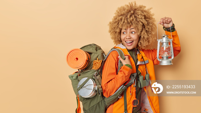 Hiking and adventure concept. Positive active female camper explores new places holds lantern to see in darkness carries backpack isolated over brown background empty space for your promotion