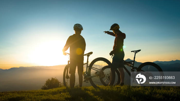 Two silhouettes standing near mountain bikes, talking and observing view in the background, mountaintops