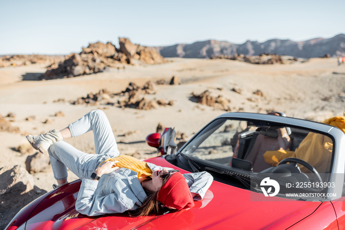 Lifestyle portrait of a young woman enjoying road trip on the desert valley, lying on the car hood and photographing on phone