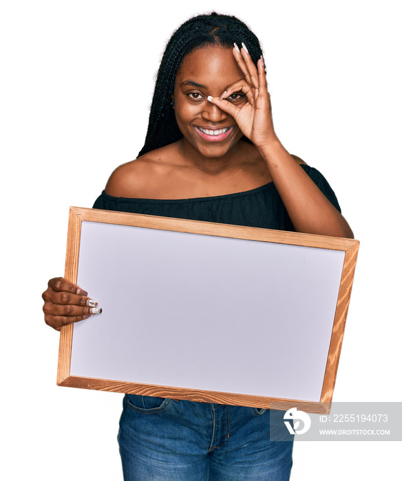 Young african american woman holding empty white chalkboard smiling happy doing ok sign with hand on eye looking through fingers