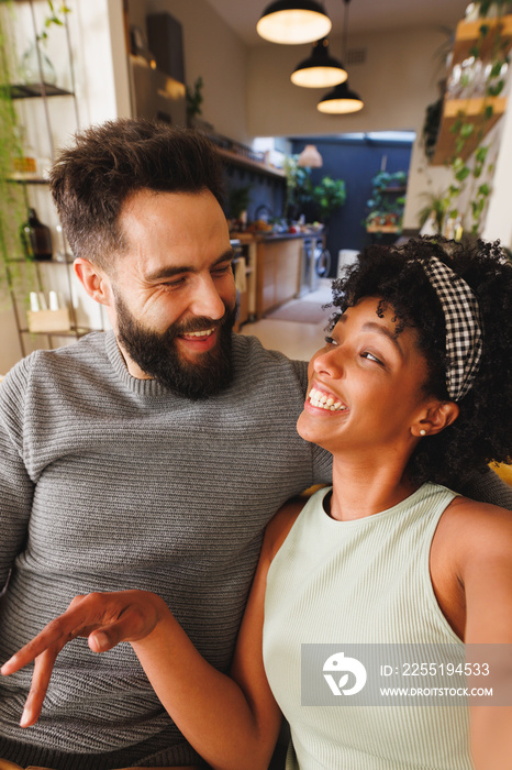 Cheerful biracial young couple looking at each other and smiling while relaxing in living room