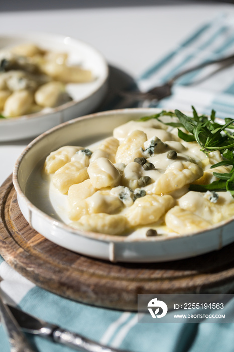 Homemade potato gnocchi with creamy gorgonzola sauce decorated with capers and arugula in white plate on wooden board.