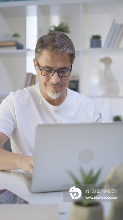 Older man in white sitting at desk in bright room working with laptop computer in home office. Mature age, middle age, mid adult casual man in 50s, confident happy smiling.