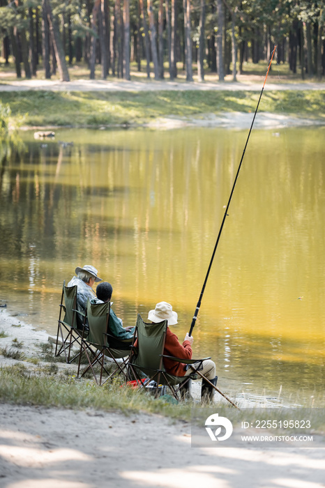 Multiethnic senior friends with fishing rod sitting near lake in park