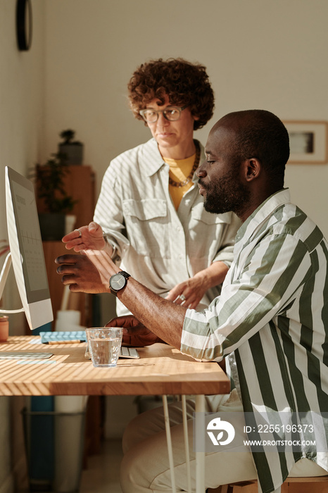 Multiethnic business couple pointing at computer monitor at table and discussing presentation together at office