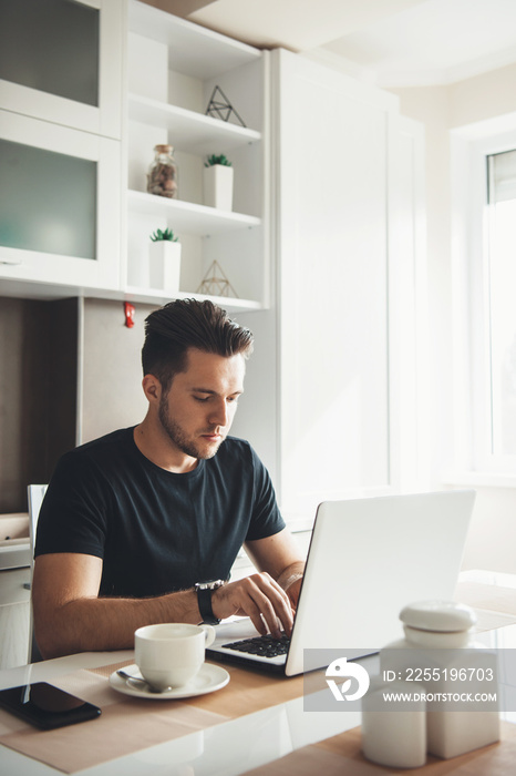 Bearded caucasian man is working remotely from home using a laptop in the kitchen while drinking a coffee