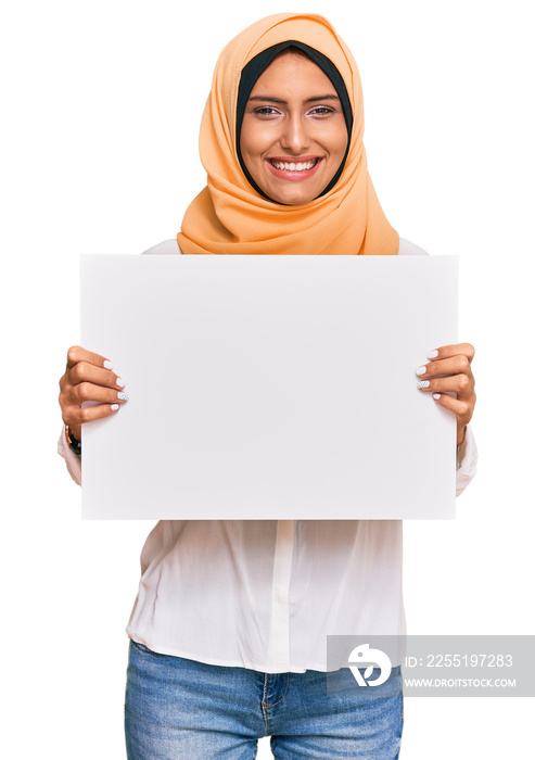 Young brunette arab woman wearing traditional islamic hijab holding banner looking positive and happy standing and smiling with a confident smile showing teeth