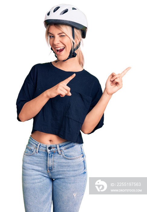 Young beautiful blonde woman wearing bike helmet smiling and looking at the camera pointing with two hands and fingers to the side.