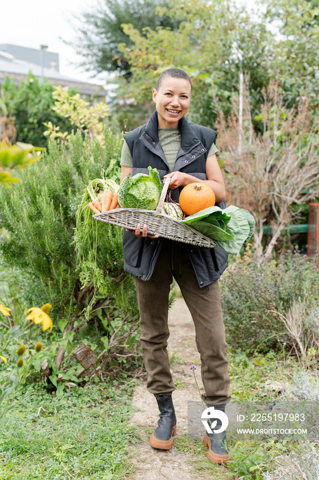 Portrait of smiling woman holding basket with fresh vegetables in urban garden