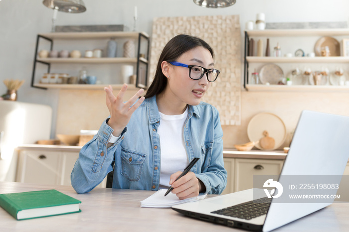 Young asian student studying at home remotely, woman watching online video course sitting in kitchen using laptop and notepad.