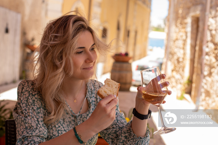 Portrait of young gorgeous woman wearing summer dress, sitting, holding glass of white wine and piece of bread toast.