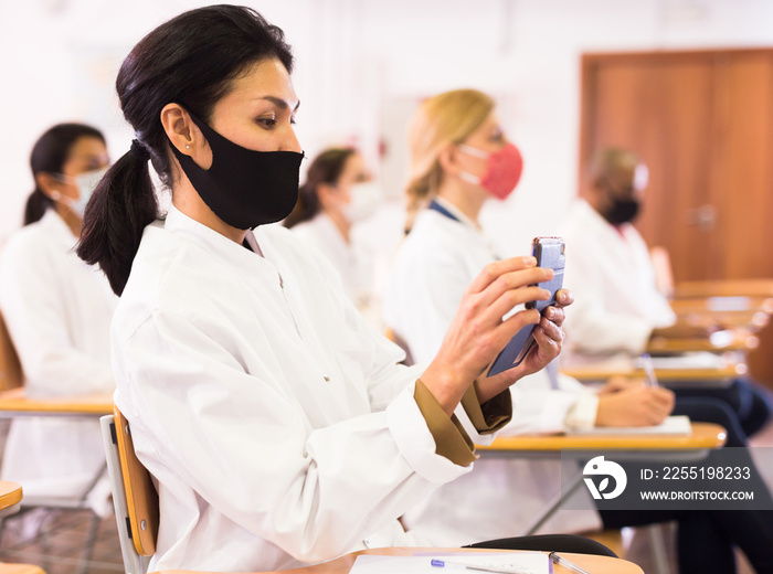 Asian female doctor in protective face mask attending medical congress, using mobile phone to record lecturer speech. New normal during coronavirus pandemic