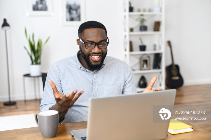 Positive and excited African-American freelancer in glasses sitting at the desk, with the laptop, working from home, feeling surprised, discussing the business project with the client on a video call