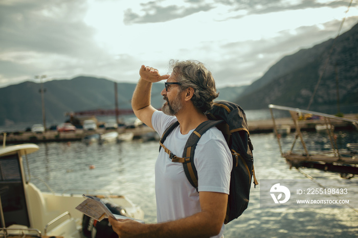 Smiling tourist mature man standing with map and backpack near the sea.