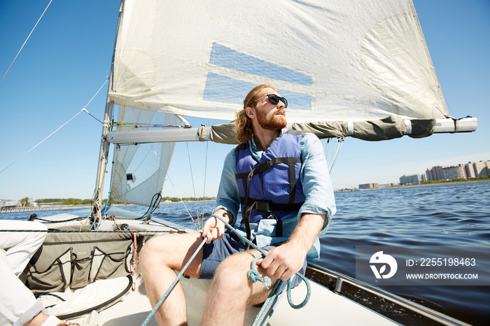 Serious pensive hipster young man with beard holding rope while operating sail boat and enjoying landscape around