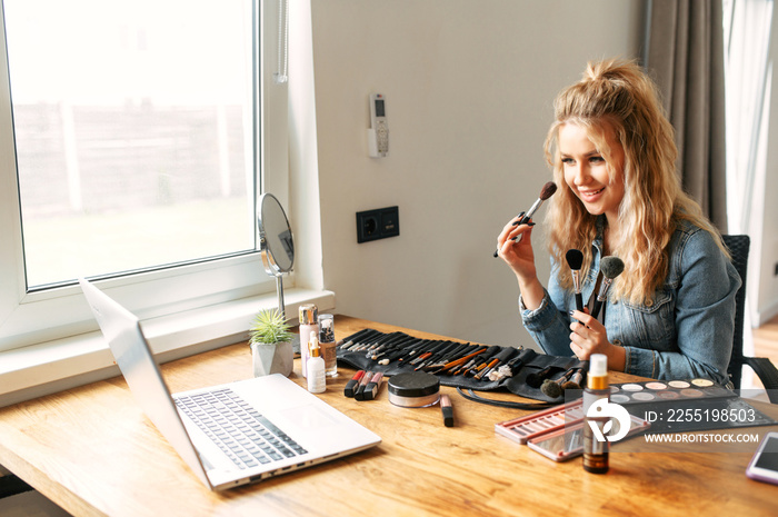 Visage teacher works from home. A young woman demonstrates how to applying powder with brushes in webcam. Online makeup classes