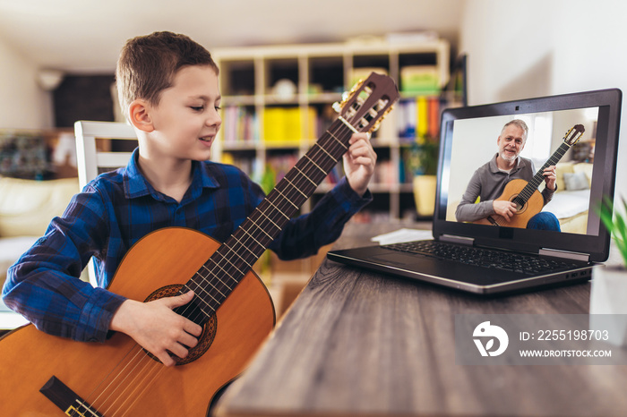 Focused boy playing acoustic guitar and watching online course on laptop while practicing at home. Online training, online classes.