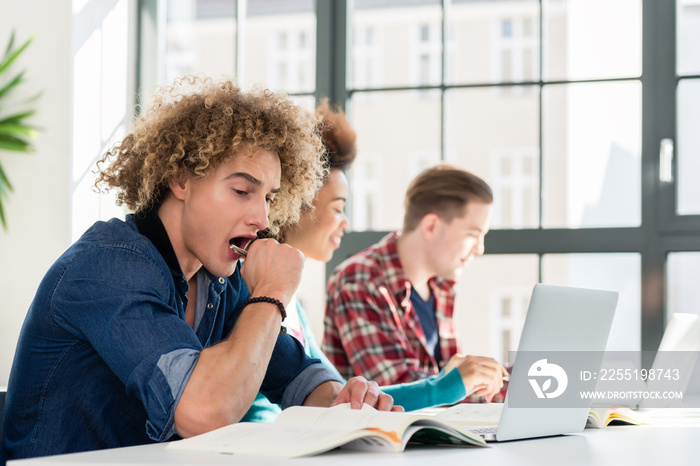 Side view portrait of a funny student yawning in front of a book, while sitting down at desk in the classroom at a modern college or university