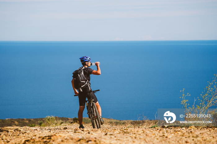 a young guy on a mountain bike stopped to drink water from a jar on a stony road near the Mediterranean Sea in Spain.