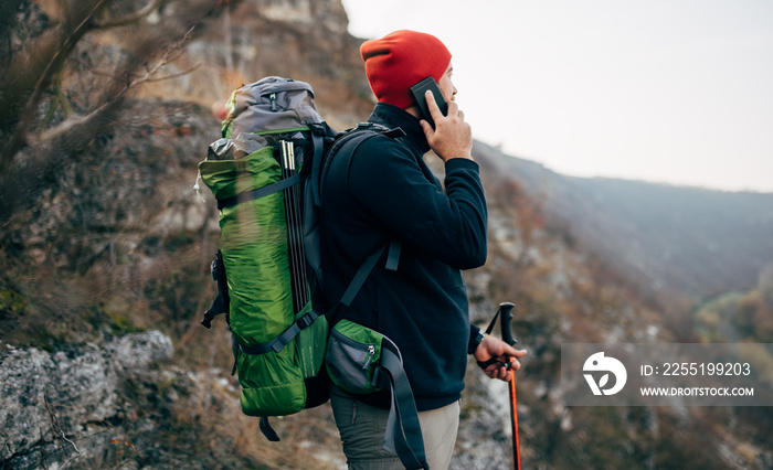 Side view portrait of young explorer man trekking in mountains, have a call with his friend. Traveler man soeaking with his family using his smart phone during hiking in moutain. Travel, people