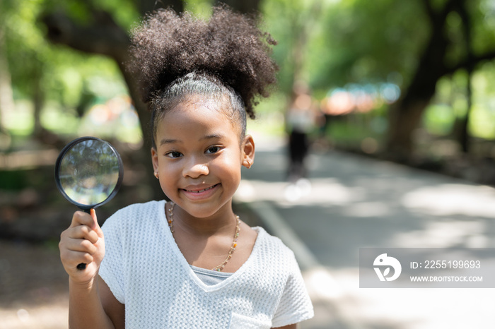 Smiling African American little girl has fun holding magnifying glass to explore and look bugs on the tree between learning beyond the classroom. Education outdoor concept.