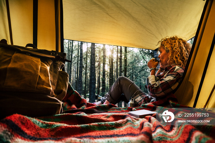 Independent beautiful curly blonde hair woman sit down outside a tent in free wild camping in the forest enjoying a cup of tea and thinking - outdoor natural lifestyle and alternative way of travel