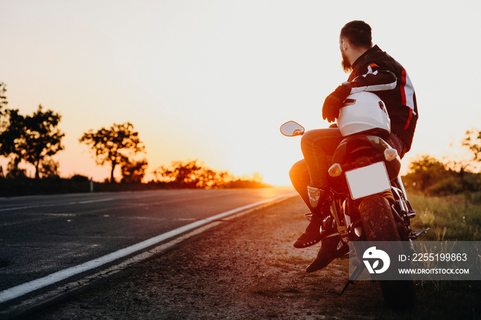 Confident attractive european biker sitting on his bike and looking at the sunset contemplating while resting in his traveling on the bike.