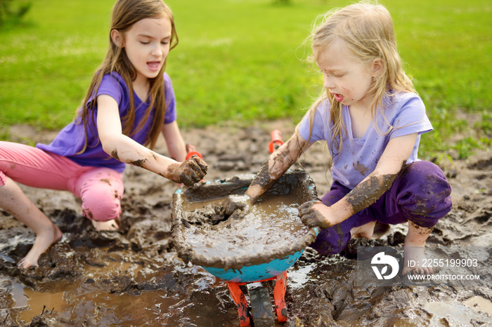 Two funny little girls playing in a large wet mud puddle on sunny summer day. Children getting dirty while digging in muddy soil.