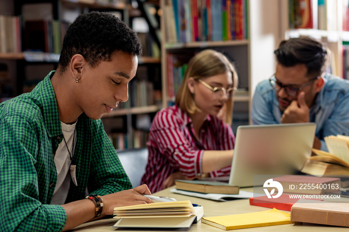 Portrait of young african american student sitting in library studying, with his friends in background.