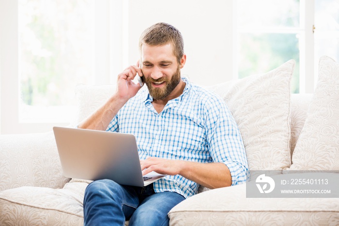 Man using laptop while talking on mobile phone in living room