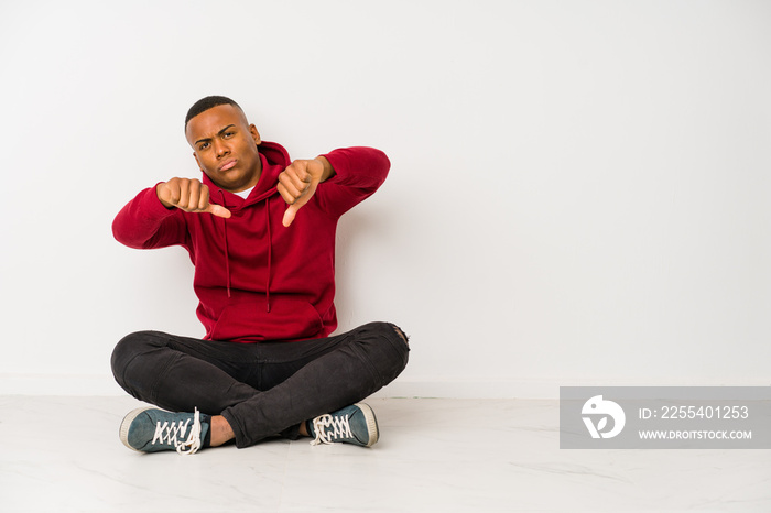 Young latin man sitting on the floor isolated showing thumb down and expressing dislike.