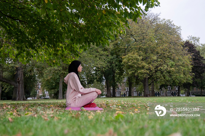 Woman in hijab and pink tracksuit meditating in park