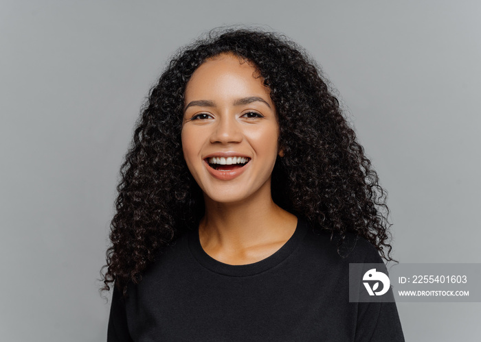 Close up shot of happy dark skinned Afro American woman laughs positively, being in good mood, dressed in black casual clothes, isolated on grey background. Human emotions and feeligs concept.