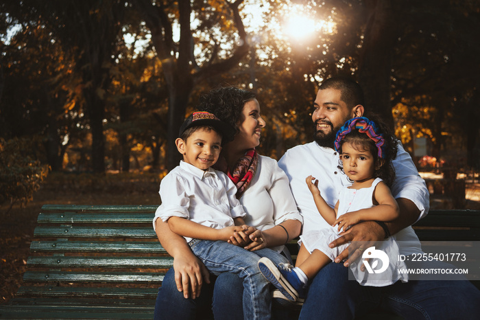 young multiracial latin and european family sitting on a park bench looking at each other with the sun behind