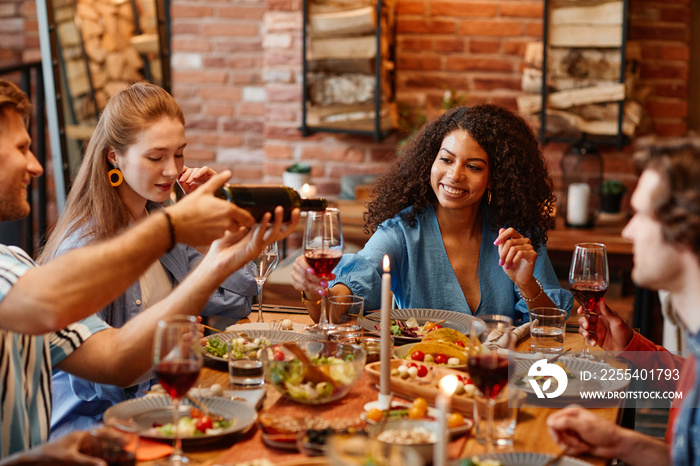 Portrait of black young woman enjoying glass of wine during dinner party with friends in cozy warm setting