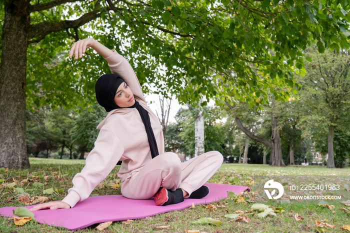 Woman in hijab and pink tracksuit practicing yoga in park