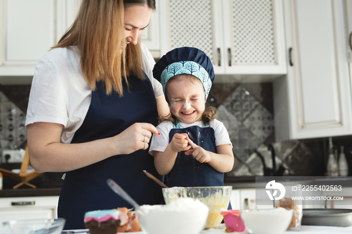 Cute little girl and her beautiful mom in matching aprons and caps play and laugh while kneading dough in the kitchen.