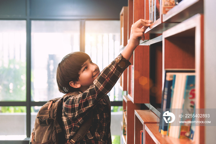 Portrait Photo of a pretty boy in red and brown plaid shirt with a gray backpack in school library or books store. Studying at the library by reading a book in free time. Self learning concept idea.