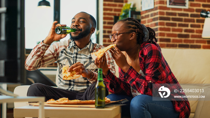 Young couple laughing and eating pizza, sitting on living room couch and enjoying fast food takeaway meal. Happy partners watching comedy movie on tv channel program. Handheld shot.