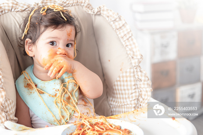 Lovely baby girl eating spaghetti and making a mess. Family leave baby alone, eating pasta herself.